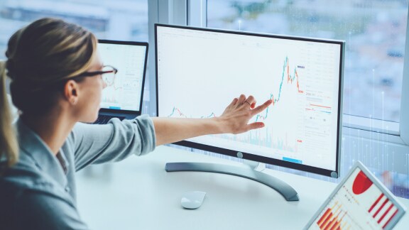 A woman pointing at a desktop monitor displaying a line graph